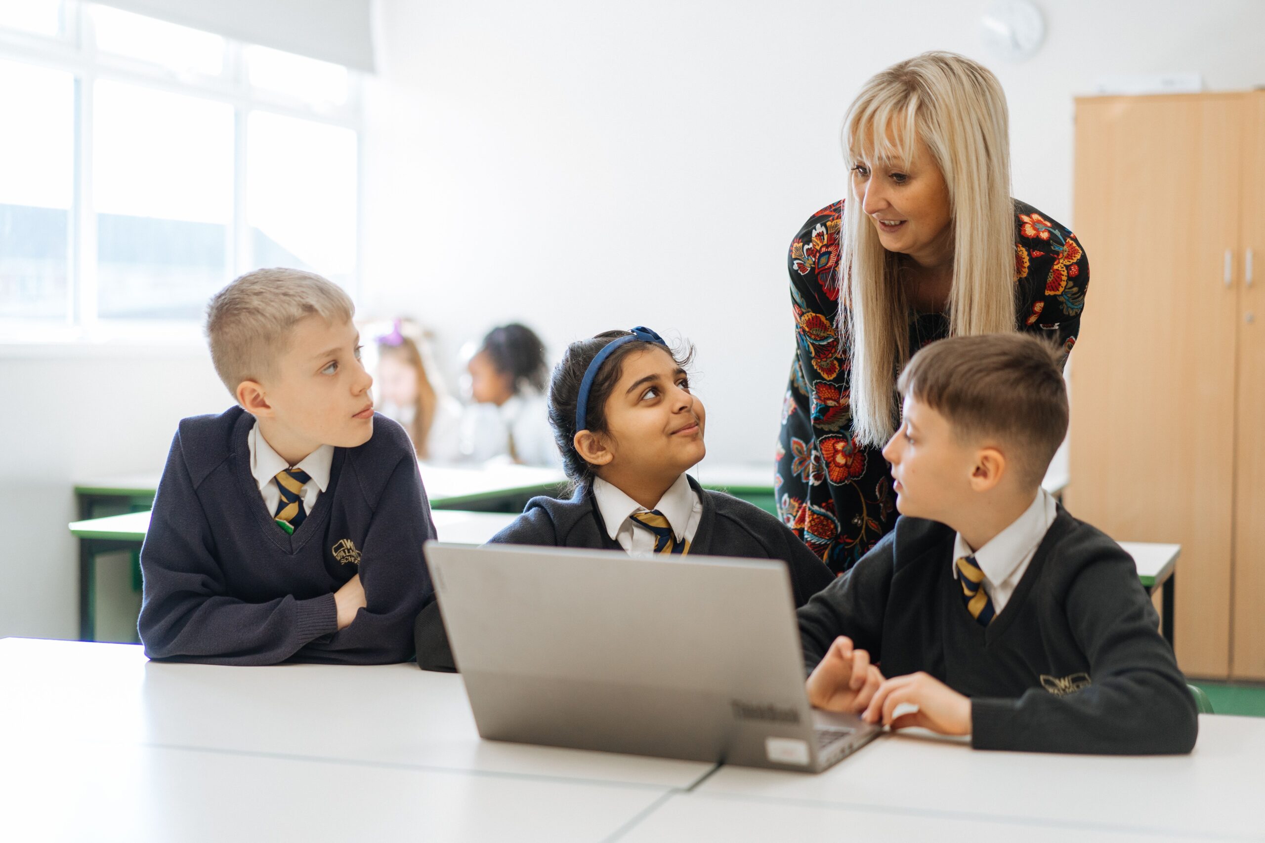 Three students sit in front of a laptop whilst their teacher stands behind them. It looks like they're discussing what is displayed on the laptop.