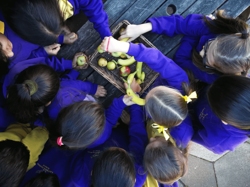 A group of around six children wearing school uniforms reach into a basket containing fruit.