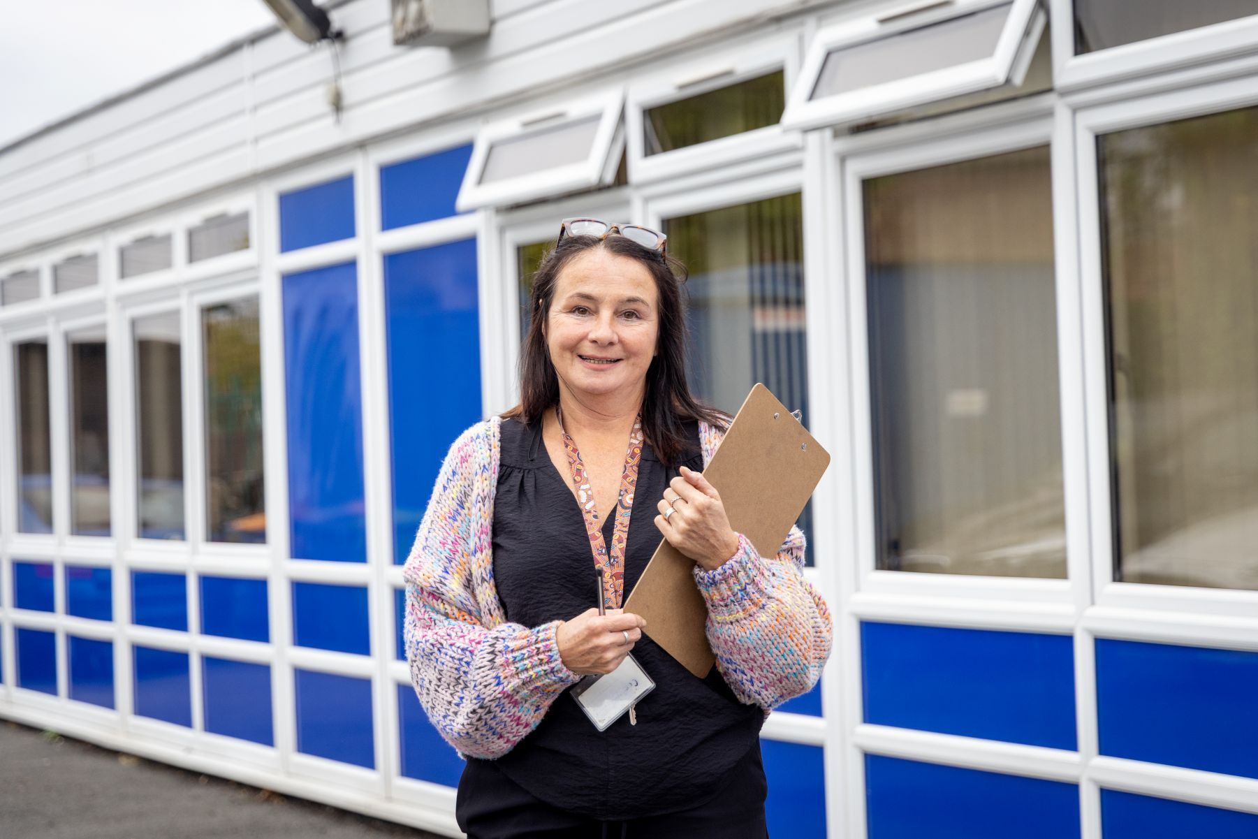 A teacher wearing a black top, multicoloured cardigan, and glasses on her head, stood outside a school building with a clipboard